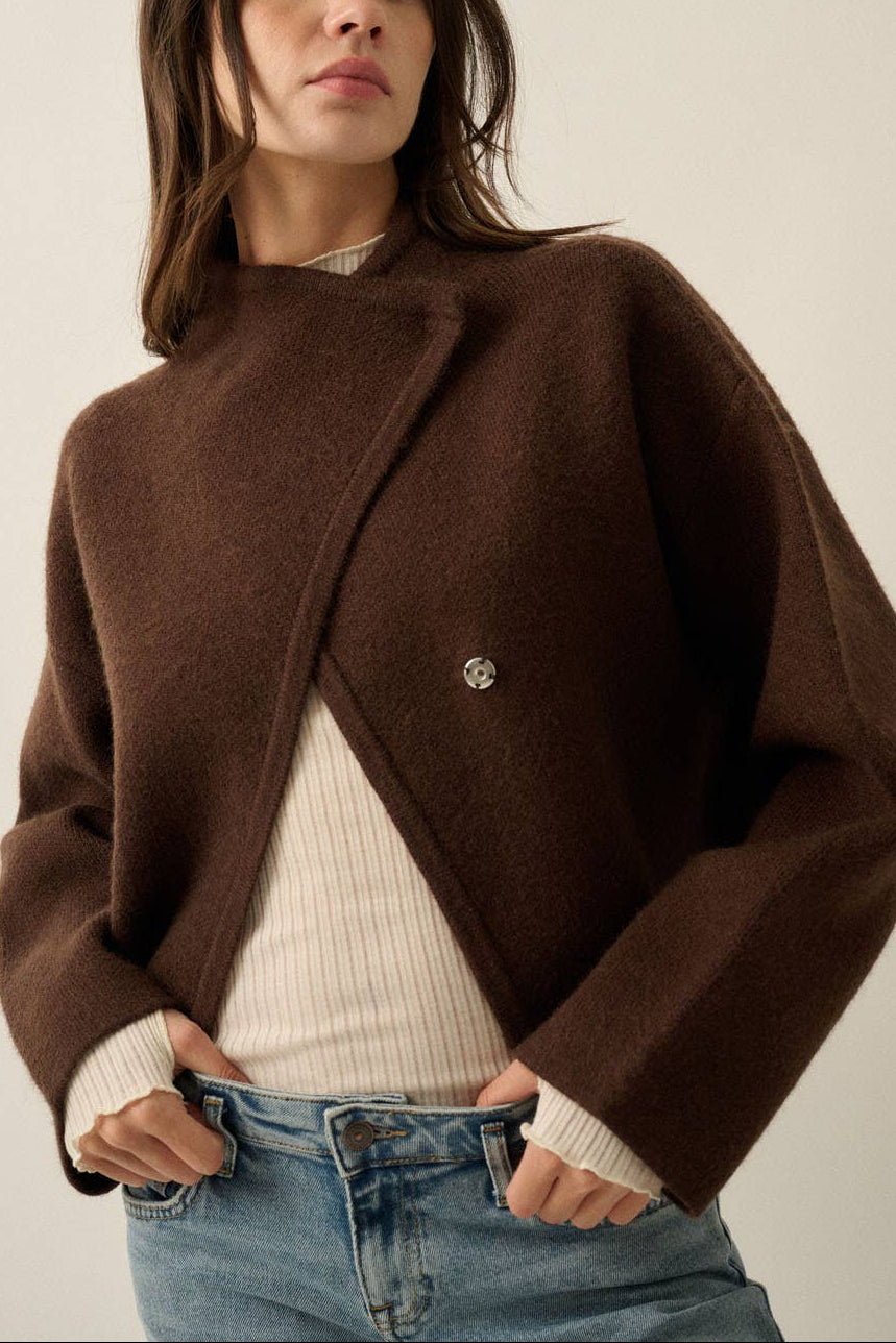 A woman wearing a dark brown cropped wrap-style coat with a single decorative button, layered over a cream ribbed sweater and blue jeans, against a plain light beige studio background.