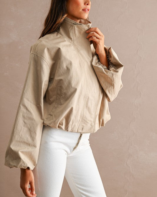 A woman wearing a cropped beige lightweight jacket with a high collar and white pants, standing against a textured beige wall.