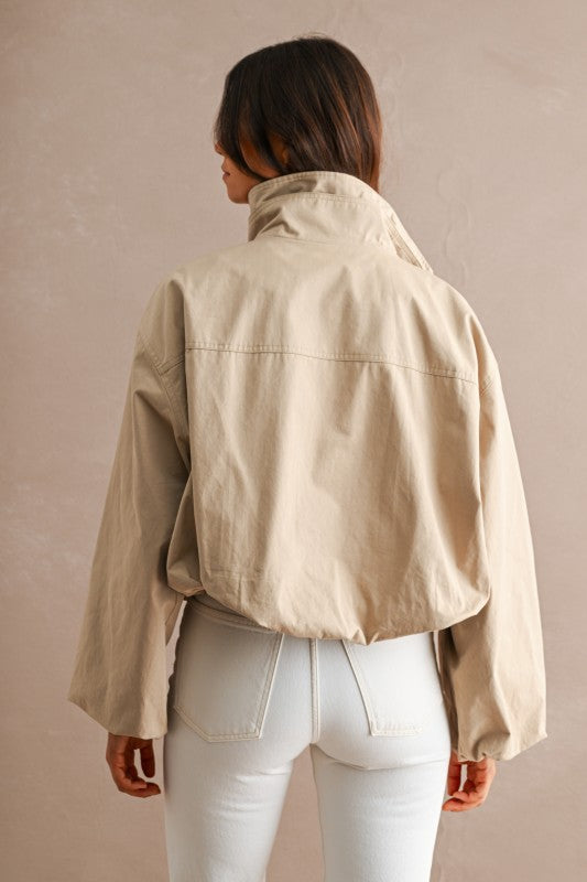 A woman shown from behind wearing a cropped beige jacket with a high collar and white pants, standing against a textured beige wall.