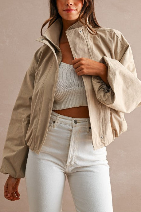 A woman modeling a light beige oversized utility jacket over a white ribbed crop top and white jeans, set against a neutral beige studio background.