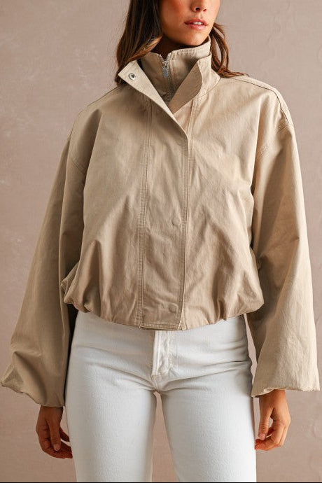 A woman wearing a beige lightweight zip-front jacket with a high stand collar and white pants, standing against a textured beige wall.