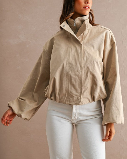 A woman wearing a beige cropped utility jacket with a high stand collar and white pants, standing against a textured beige wall.