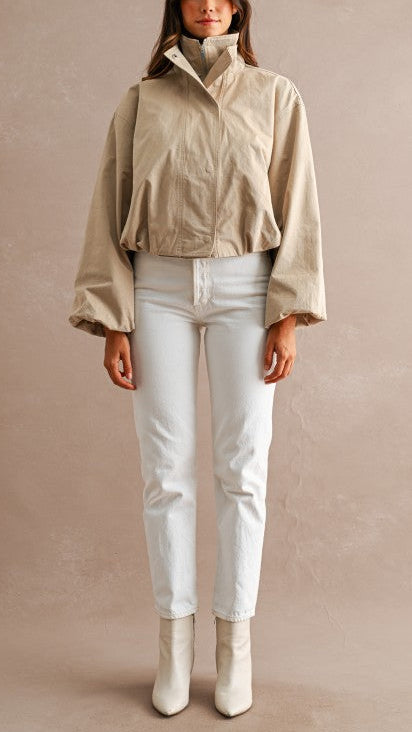 A woman stands against a textured beige studio backdrop wearing a cropped beige jacket with a high stand collar, white pants, and off-white ankle boots.