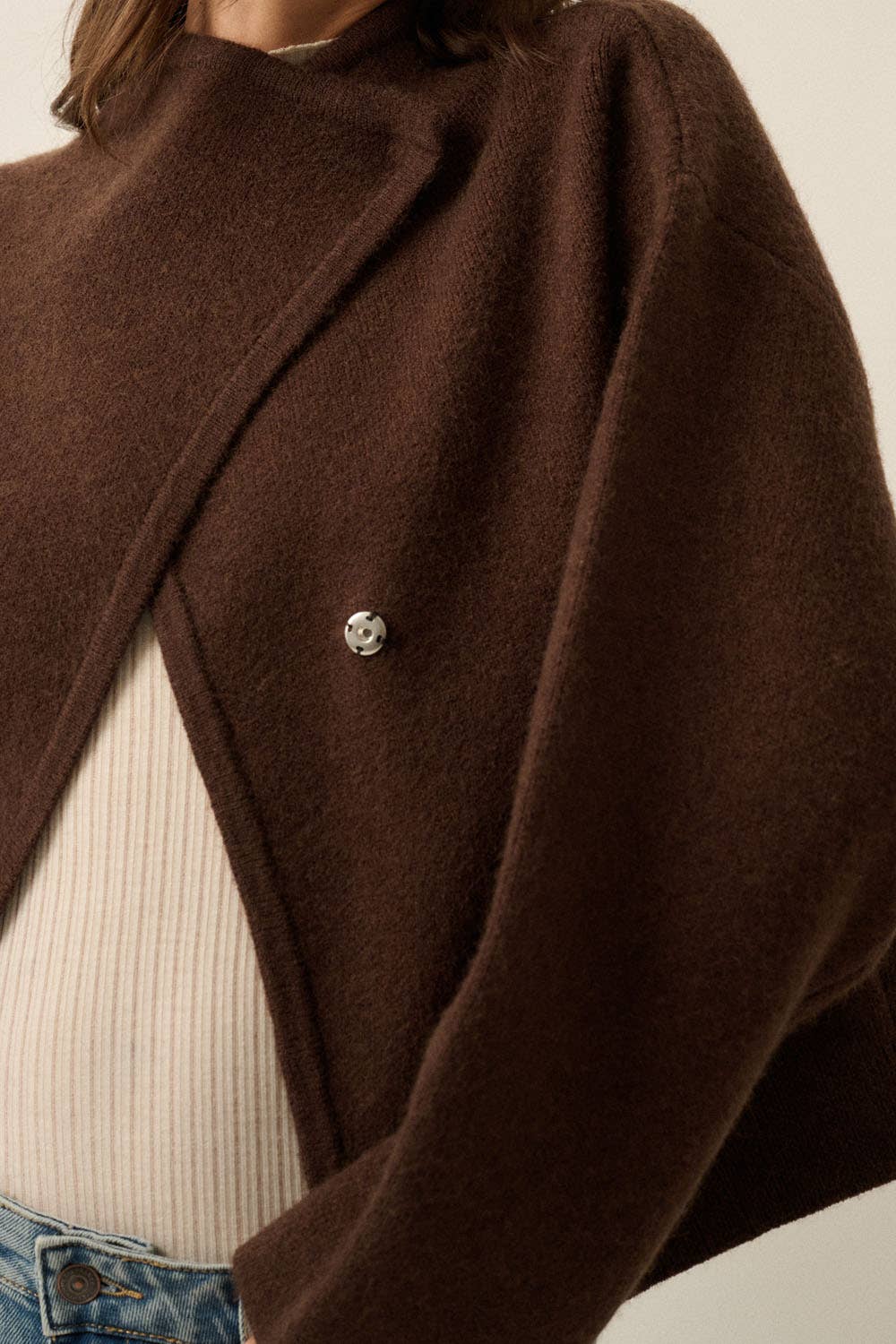A close-up of a person wearing a dark brown cropped cardigan with a silver button, layered over a cream ribbed top and blue jeans.