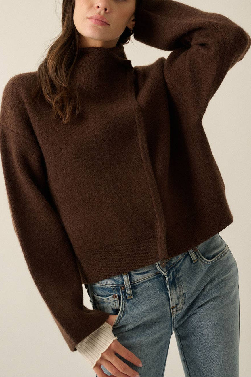 A woman wearing a dark brown cropped knit sweater with a mock neck and wide sleeves, paired with light blue jeans, photographed against a plain light beige studio background.