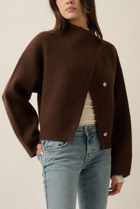 A woman modeling a cropped brown knit cardigan with wide sleeves, paired with light-wash blue flare jeans and dark brown shoes, against a plain beige studio background.