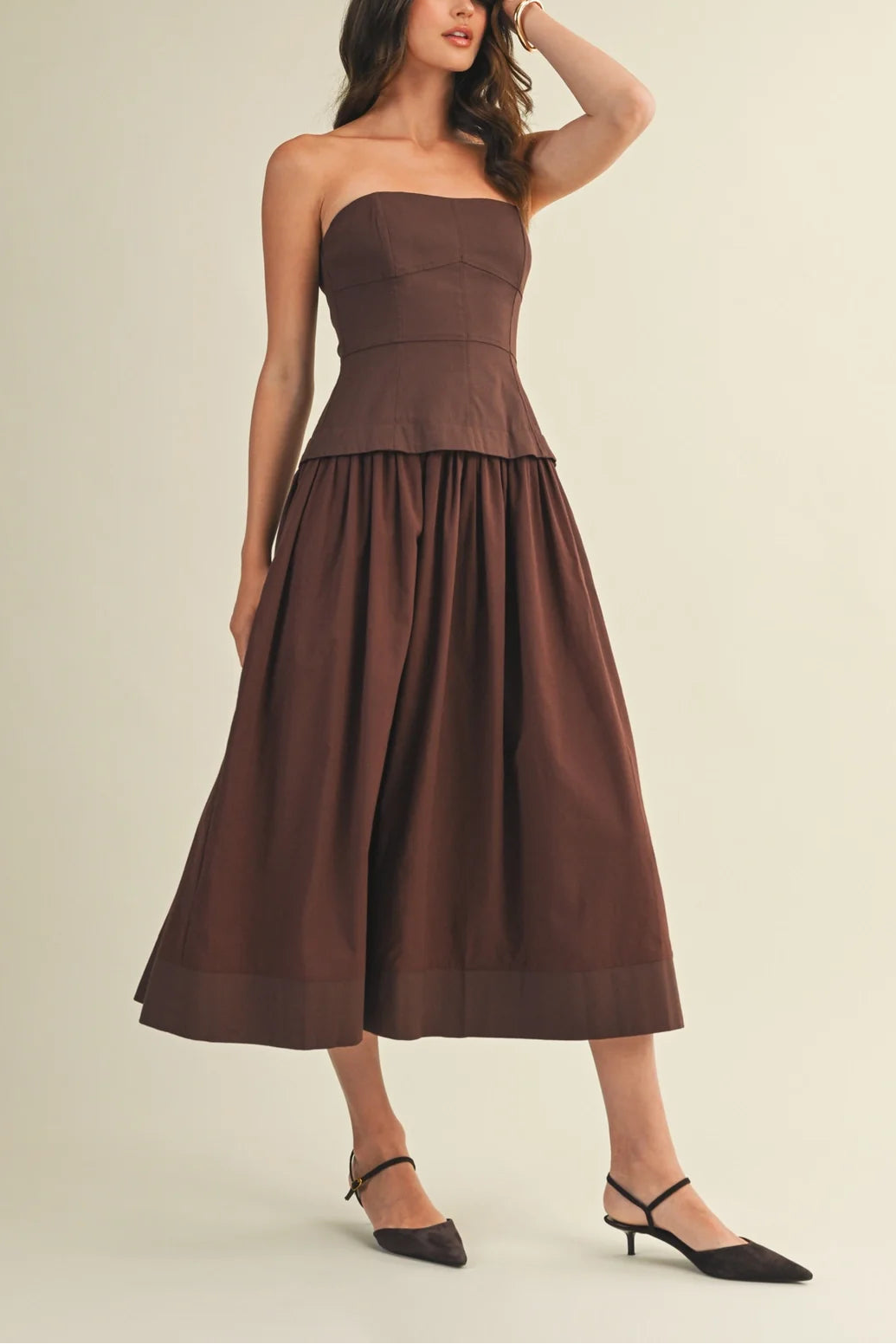 A woman models a strapless brown midi dress with a structured bodice and a full gathered skirt, paired with black pointed-toe heels, against a plain light beige studio background.