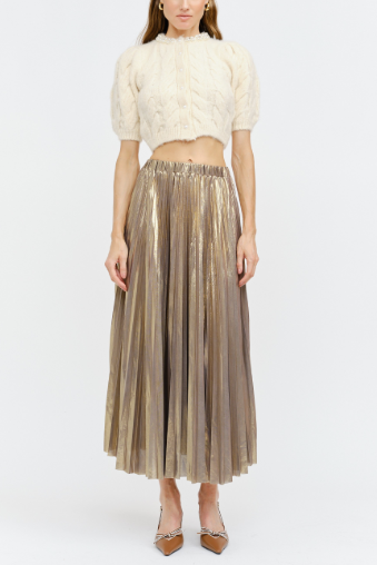 A model stands against a light gray studio background wearing a cream-colored short-sleeve knit crop top and a metallic champagne-gold pleated midi skirt, paired with brown pointed-toe heels.