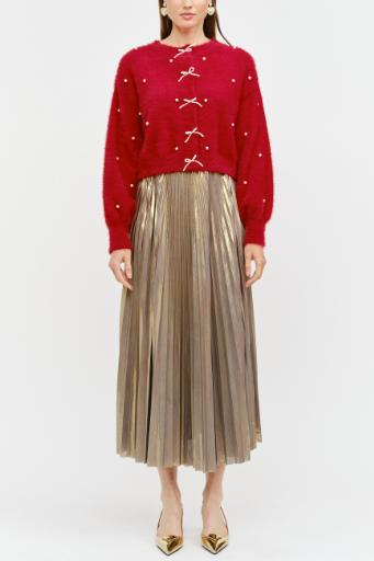 A model stands against a light gray studio background wearing a bright red cropped knit sweater with decorative silver bow details down the front, paired with a metallic champagne pleated midi skirt and pointed gold heels.