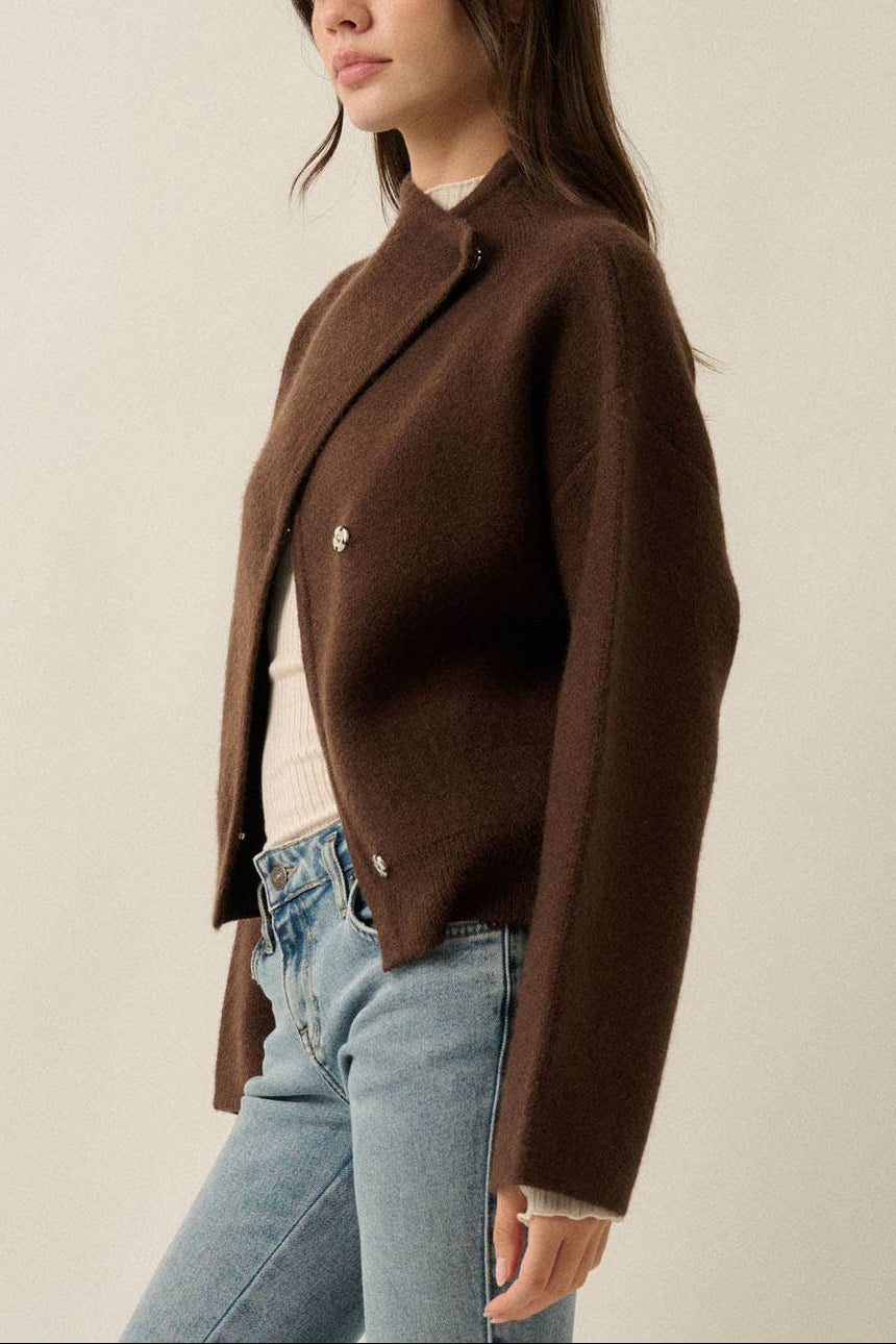 A woman wearing a brown cropped cardigan with a high collar and silver-tone buttons, paired with light blue jeans, photographed against a plain light beige studio background.