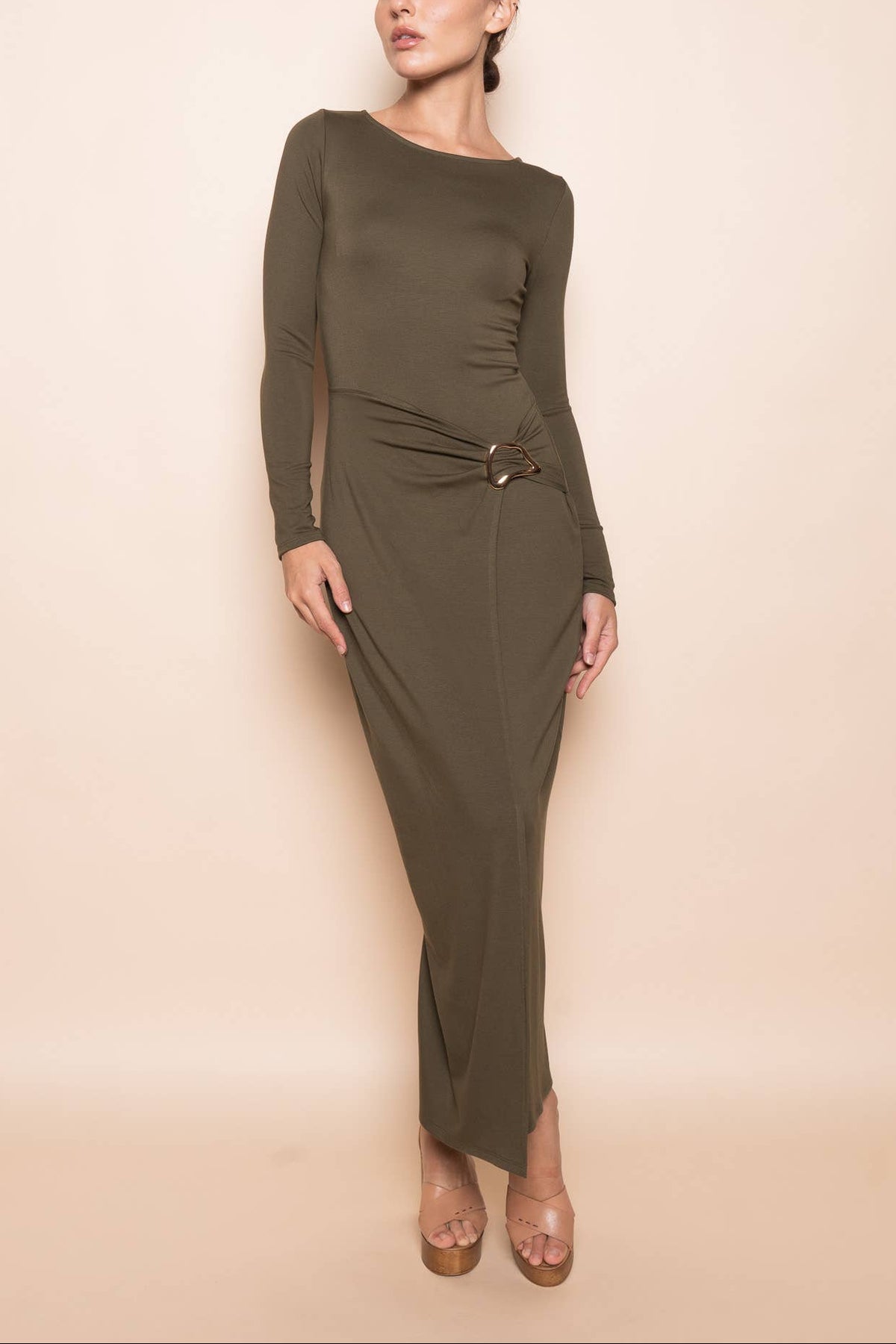 A woman modeling a long-sleeve, olive green maxi dress with a ruched waist and a small metallic ring detail, set against a solid beige studio background.
