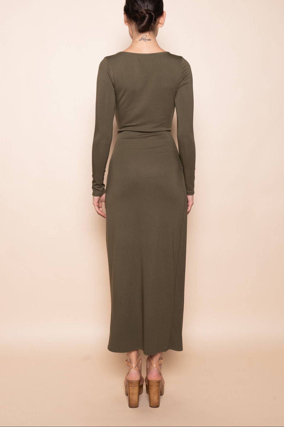 A woman viewed from the back wearing a long-sleeve olive green maxi dress and brown block-heel sandals against a solid beige studio background.