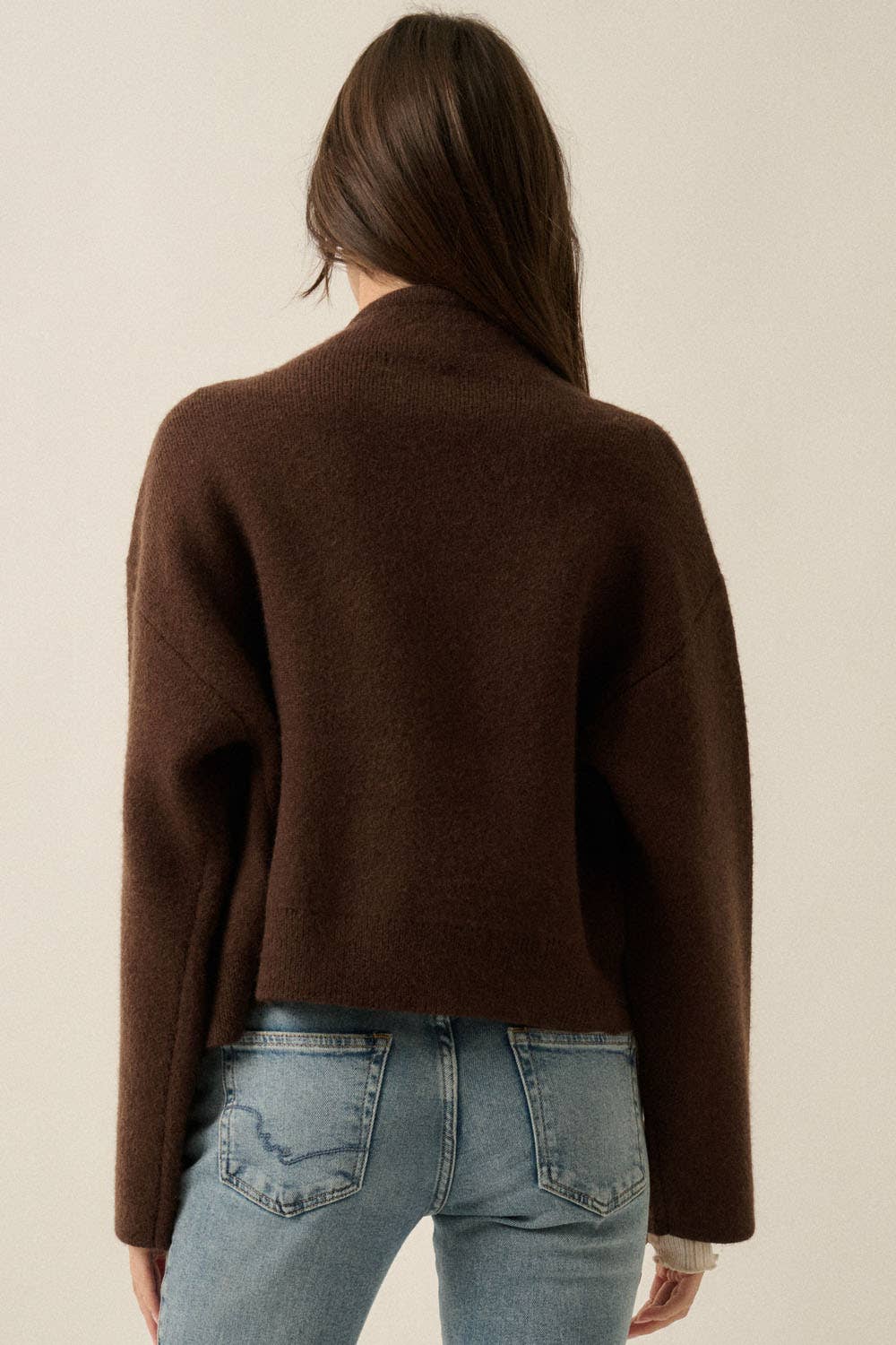 A woman viewed from the back wearing a dark brown cropped knit sweater and light blue jeans against a plain light beige studio background.